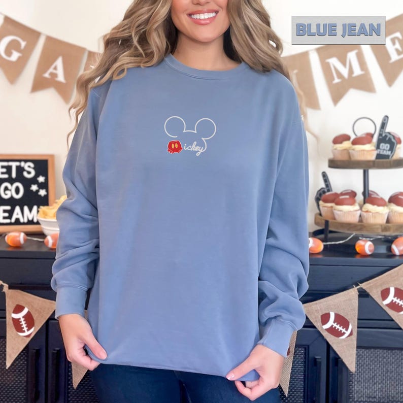 Peut inclure: Sweat-shirt bleu jean avec un contour de Mickey Mouse et le mot "mickey" en &eacute;criture manuscrite. Le contour de Mickey Mouse est blanc avec un n&oelig;ud papillon rouge et jaune. Le texte "BLUE JEAN" est en haut de l'image.