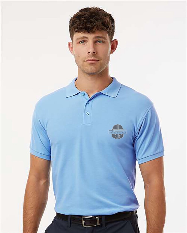 K&ouml;nnte beinhalten: Hellblaues Poloshirt mit kurzen &Auml;rmeln und Kragen. Das Shirt hat eine Knopfleiste mit drei Kn&ouml;pfen und ein kleines Logo auf der linken Brust. Das Logo enth&auml;lt den Text "THIS IS THE WAY".