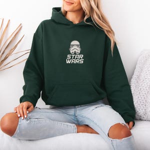 May include: A dark green hooded sweatshirt with a front pocket and the text "STAR WARS" in white. The sweatshirt features a white graphic of a Stormtrooper helmet. The model is wearing light blue jeans.
