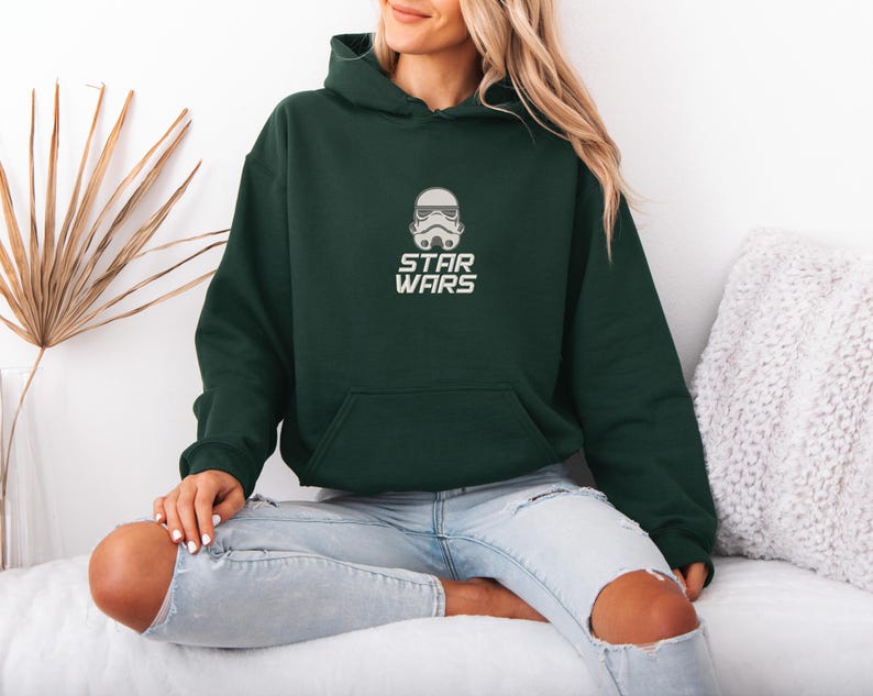 May include: A dark green hooded sweatshirt with a front pocket and the text "STAR WARS" in white. The sweatshirt features a white graphic of a Stormtrooper helmet. The model is wearing light blue jeans.