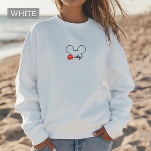 Peut inclure: Sweat-shirt blanc avec un motif minimaliste de Mickey Mouse. Le motif comprend un contour noir de la t&ecirc;te de Mickey, une bouche rouge et le mot "Mickey" dans une police manuscrite. Le mot "WHITE" est dans un encadr&eacute; blanc en haut &agrave; gauche.