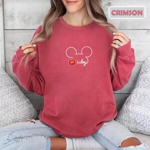 Peut inclure: Sweat-shirt de couleur cramoisie avec un contour blanc des oreilles de Mickey Mouse et le mot "Mickey" en cursive. Le motif comprend une petite t&ecirc;te de Mickey Mouse rouge, noire et blanche. Le sweat-shirt est de couleur unie.