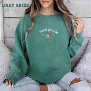 May include: Light green sweatshirt with the text "DisneyWorld" in a script font, with "EST. 1971" below. The sweatshirt features a graphic of Mickey Mouse wearing a Santa hat. The sweatshirt is a solid light green colour.