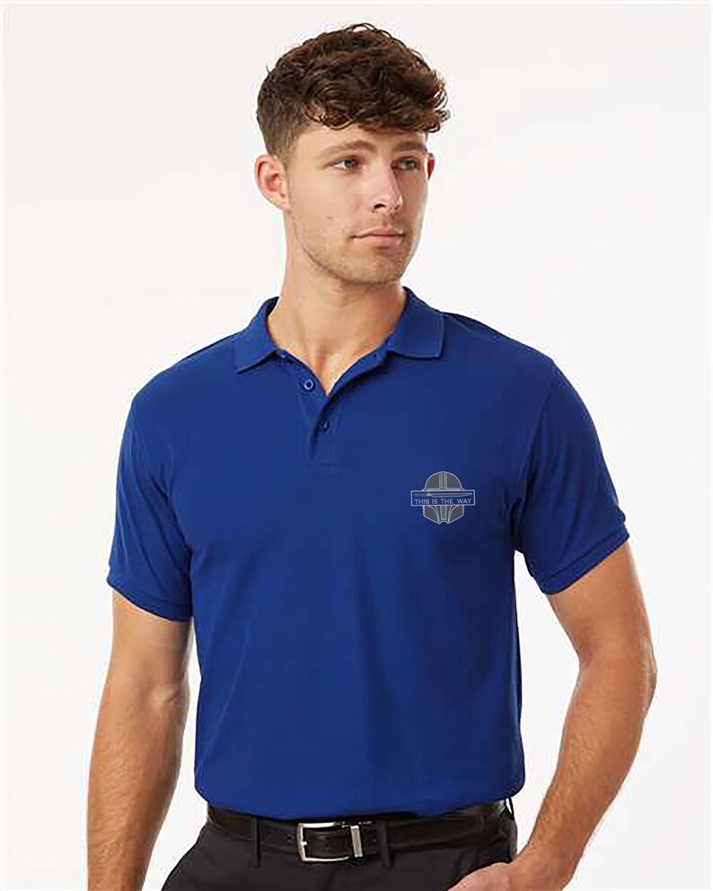 K&ouml;nnte beinhalten: Ein kurz&auml;rmeliges, k&ouml;nigsblaues Poloshirt mit Kragen und Dreiknopfleiste. Das Shirt hat ein kleines, hellblaues Emblem auf der linken Brust und den Text "THIS IS THE WAY". Das Model tr&auml;gt einen schwarzen G&uuml;rtel und eine schwarze Hose.