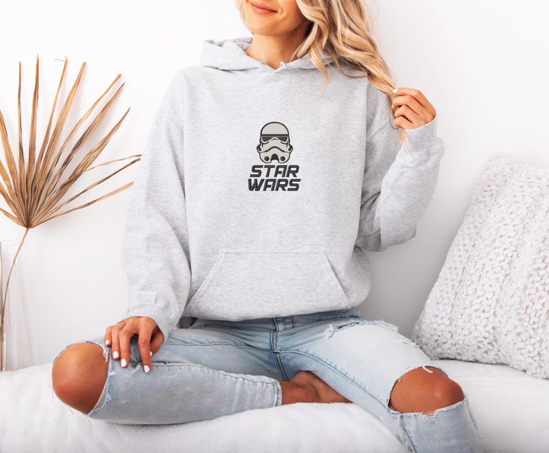 May include: A light gray hoodie with a front pocket and a drawstring hood. The hoodie features a small graphic of a Stormtrooper helmet and the words "STAR WARS" in black. The model is wearing light blue jeans.