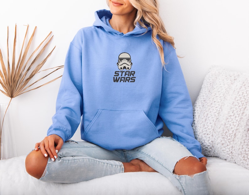 May include: A light blue hoodie with a front pocket and a hood. The hoodie features a black and white graphic of a Stormtrooper helmet and the words "STAR WARS" in black. The person is wearing light blue jeans.