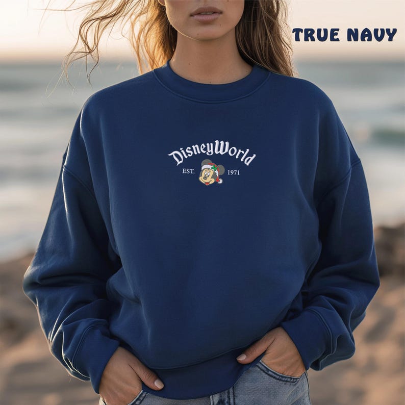May include: Navy blue sweatshirt featuring the text "DisneyWorld" in white script, with a small Mickey Mouse graphic wearing a Santa hat. The text "EST. 1971" is also included. The words "TRUE NAVY" are printed in the upper right corner of the sweatshirt.