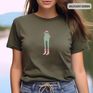 May include: Military green t-shirt featuring a cartoon frog wearing a cowboy hat and pink boots with hearts. The frog is standing with its hands on its hips. The text "MILITARY GREEN" is in a white rectangle in the upper right corner.