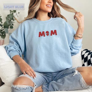 Pode incluir: Camisola azul clara com a palavra "MOM" em vermelho, com um design inspirado no Homem-Aranha. A camisola de manga comprida e gola redonda tem o texto "LIGHT BLUE" numa caixa no canto superior esquerdo.