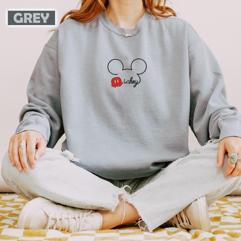 Peut inclure: Un sweat-shirt gris clair avec un contour noir de la t&ecirc;te de Mickey Mouse et le mot "Mickey" en script noir. Le design comprend une bouche rouge. Le mot "GREY" est dans un rectangle gris en haut &agrave; gauche.