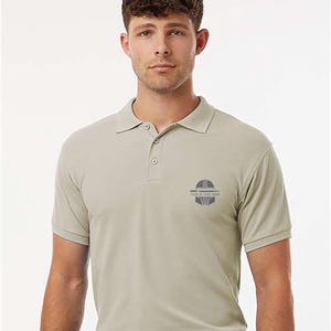 K&ouml;nnte beinhalten: Hellbeige Polo-Shirt mit kurzen &Auml;rmeln und Kragen. Das Shirt hat eine Knopfleiste mit drei Kn&ouml;pfen und eine kleine Grafik auf der linken Brust mit dem Text "THIS IS THE WAY". Die Person tr&auml;gt einen schwarzen G&uuml;rtel und eine marineblaue Hose.