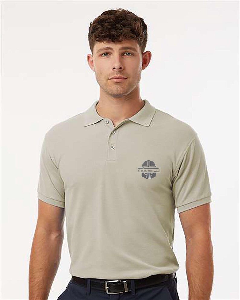 K&ouml;nnte beinhalten: Hellbeige Polo-Shirt mit kurzen &Auml;rmeln und Kragen. Das Shirt hat eine Knopfleiste mit drei Kn&ouml;pfen und eine kleine Grafik auf der linken Brust mit dem Text "THIS IS THE WAY". Die Person tr&auml;gt einen schwarzen G&uuml;rtel und eine marineblaue Hose.