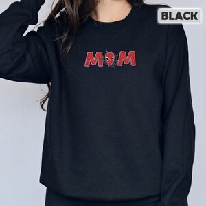 Pode incluir: Camisola preta com a palavra "MOM" em vermelho, estilizada com um design de teia de aranha e uma m&aacute;scara do Homem-Aranha. A palavra "BLACK" est&aacute; numa caixa branca no canto superior direito. A camisola &eacute; de cor preta lisa.