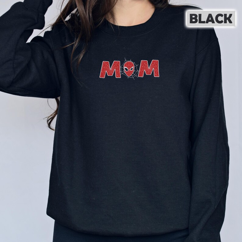 Pode incluir: Camisola preta com a palavra "MOM" em vermelho, estilizada com um design de teia de aranha e uma m&aacute;scara do Homem-Aranha. A palavra "BLACK" est&aacute; numa caixa branca no canto superior direito. A camisola &eacute; de cor preta lisa.