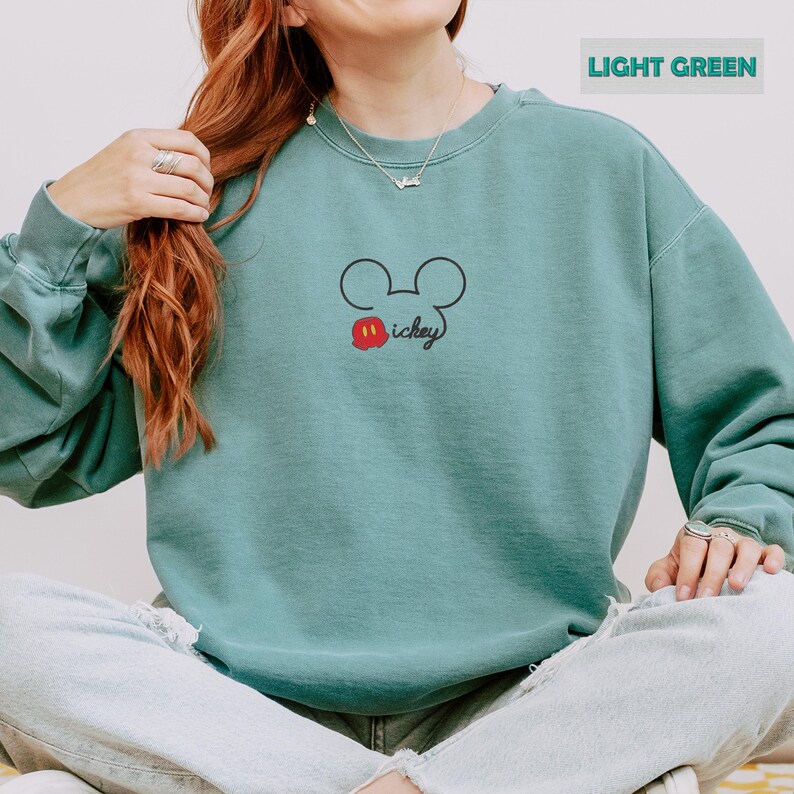 Peut inclure: Sweat-shirt vert clair avec un contour noir de la t&ecirc;te de Mickey Mouse, une bouche rouge et blanche et le mot "Mickey". Le sweat-shirt est de couleur vert clair uni.