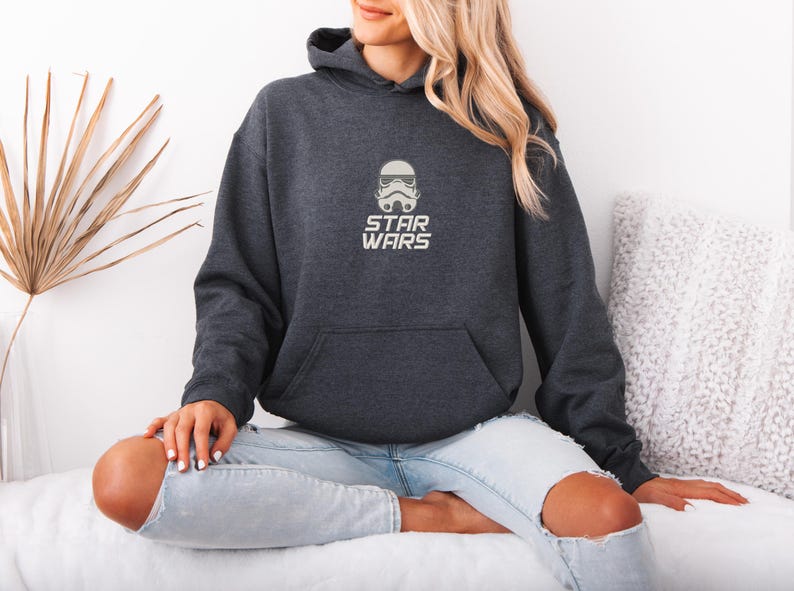 May include: Dark gray Star Wars hoodie with a white Stormtrooper helmet graphic and the words "STAR WARS" below it. The hoodie has a front pocket and a hood. The person is wearing light blue jeans.