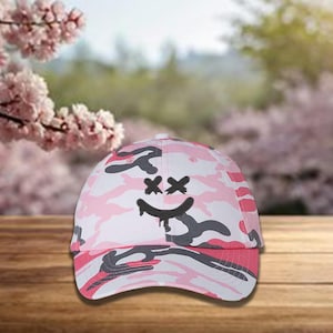 May include: A pink, white, and gray camouflage baseball cap with a curved brim. The cap features a black smiley face design with crossed-out eyes and a dripping mouth. The hat is made of a durable fabric and is suitable for casual wear.