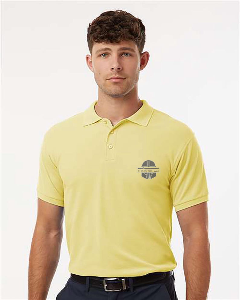 K&ouml;nnte beinhalten: Hellgelbes Poloshirt mit kurzen &Auml;rmeln und Kragen. Auf der Brust befindet sich eine kleine Grafik mit dem Text "THIS IS THE WAY". Das Shirt wird mit einer dunkelblauen Hose und einem schwarzen G&uuml;rtel getragen.