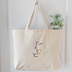 May include: A beige canvas tote bag with a black, red, and yellow outline of a cartoon mouse. The bag is hanging on a white door. A small green plant is visible inside the bag.