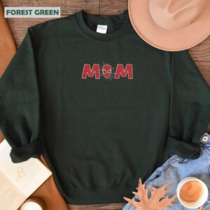 Pode incluir: Camisola verde floresta com a palavra "MOM" em vermelho, com um design de teia de aranha. A camisola &eacute; de gola redonda. O texto "FOREST GREEN" est&aacute; no canto superior esquerdo.
