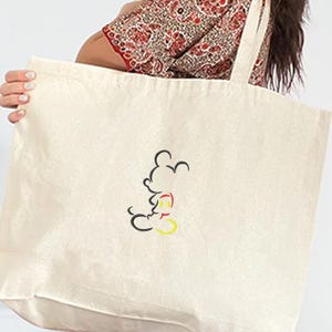 May include: Cream-colored tote bag with a black, red, and yellow outline of Mickey Mouse. The bag has long straps and is made of a sturdy fabric, suitable for carrying groceries or other items. The design is simple and classic.