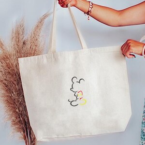 May include: Cream-colored tote bag with a black, red, and yellow Mickey Mouse outline design. The bag has long, cream-colored handles and is held up by a person. The bag is made of a sturdy canvas material.