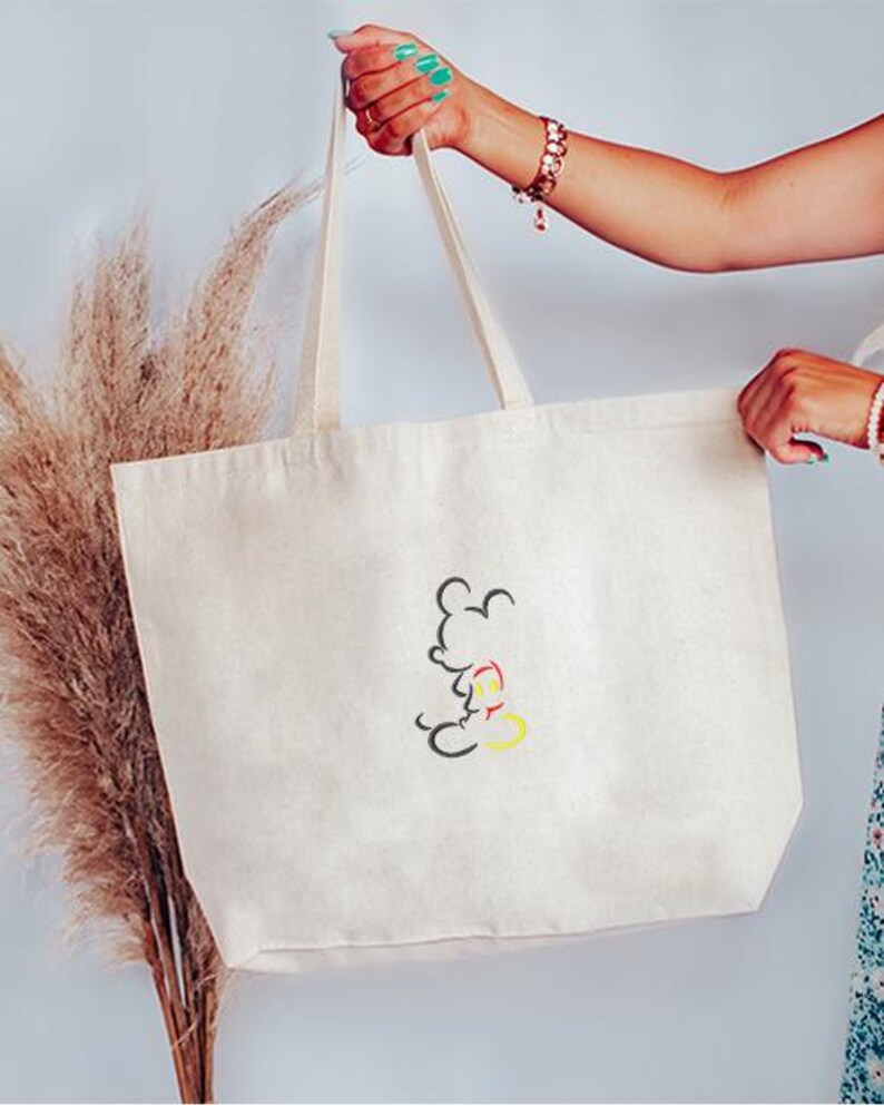 May include: Cream-colored tote bag with a black, red, and yellow Mickey Mouse outline design. The bag has long, cream-colored handles and is held up by a person. The bag is made of a sturdy canvas material.