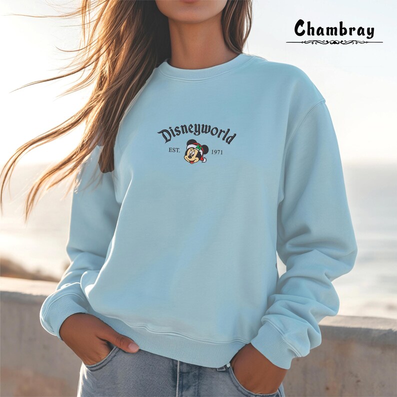 May include: A pale blue sweatshirt with the text "Disneyworld" and a Mickey Mouse graphic wearing a Santa hat. The text "EST. 1971" is below the graphic. The brand name "Chambray" is at the top right.