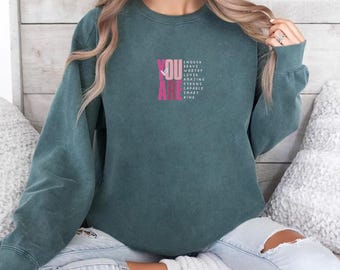 May include: A teal blue sweatshirt with the words "YOU ARE" in pink, with a list of positive affirmations below. The words "BLUE SPRUCE" are printed in a light color at the top right of the image. The sweatshirt is worn by a person.