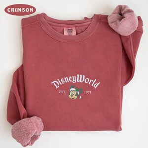 May include: Crimson sweatshirt featuring "DisneyWorld" in white script, with a Mickey Mouse graphic and "EST. 1971". The crew neck sweatshirt has ribbed cuffs. The colour is a deep red.