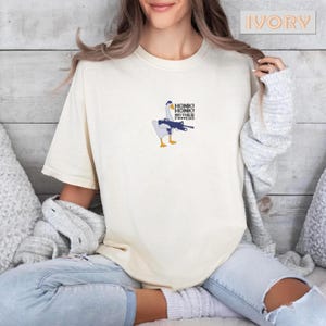 May include: Ivory-colored t-shirt featuring a cartoon goose holding a machine gun. The goose is white, blue, and yellow, with the text "HONK! HONK! MOTHER F***ERS" above it. The shirt is a relaxed fit.