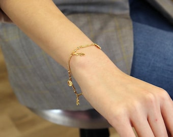 Pode incluir: Um bracelete de corrente dourada com pequenos pingentes, incluindo folhas e círculos. O bracelete está no pulso de uma pessoa.