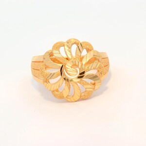 May include: A gold ring featuring a large, circular floral motif composed of interconnected, curved petals. The surface of the gold displays a combination of polished, reflective diagonal stripes and a textured, matte finish throughout the design.