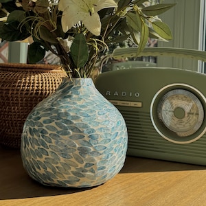 May include: A still life arrangement featuring a blue and white mosaic vase holding white lilies and greenery. A vintage green radio sits to the right. A woven basket is partially visible on the left. The scene is set on a wooden surface.