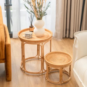 May include: Two round woven rattan side tables with a white ceramic vase with dried flowers on the larger table. The tables are on a wooden floor.