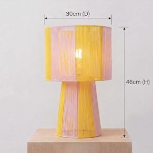 May include: A decorative table lamp with a yellow and pink striped shade and base. The lamp is 46cm (18 inches) tall and 30cm (12 inches) in diameter. The lamp is made of woven material and sits on a wooden surface.