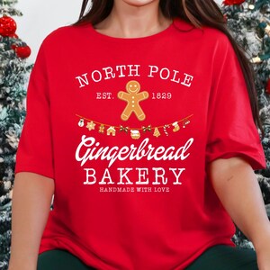 May include: A red t-shirt with the text "North Pole Gingerbread Bakery" in white. The shirt features a gingerbread man graphic and is paired with green sweatpants and festive Christmas decor.