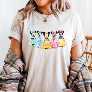 May include: A beige t-shirt featuring a colorful graphic of five Minnie Mouse characters in different dresses. Each Minnie Mouse is wearing a different colored dress, including teal, blue, yellow, pink, and yellow. The shirt is worn by a person.
