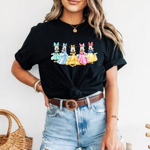May include: Black t-shirt featuring a colorful graphic of five cartoon characters in dresses. The characters are in various colors, including blue, yellow, and pink. The shirt is a classic crew neck style.