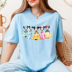 May include: Light blue t-shirt featuring a graphic of five Minnie Mouse characters in colorful dresses. The cartoon characters are arranged in a row, each wearing a different colored dress and bow. The shirt is made of a soft material.