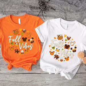 May include: Two t-shirts, one orange and one white, feature a circular design with the text "Fall Vibes." The design includes pumpkins, Mickey Mouse silhouettes, and fall-themed graphics. The shirts are short-sleeved.