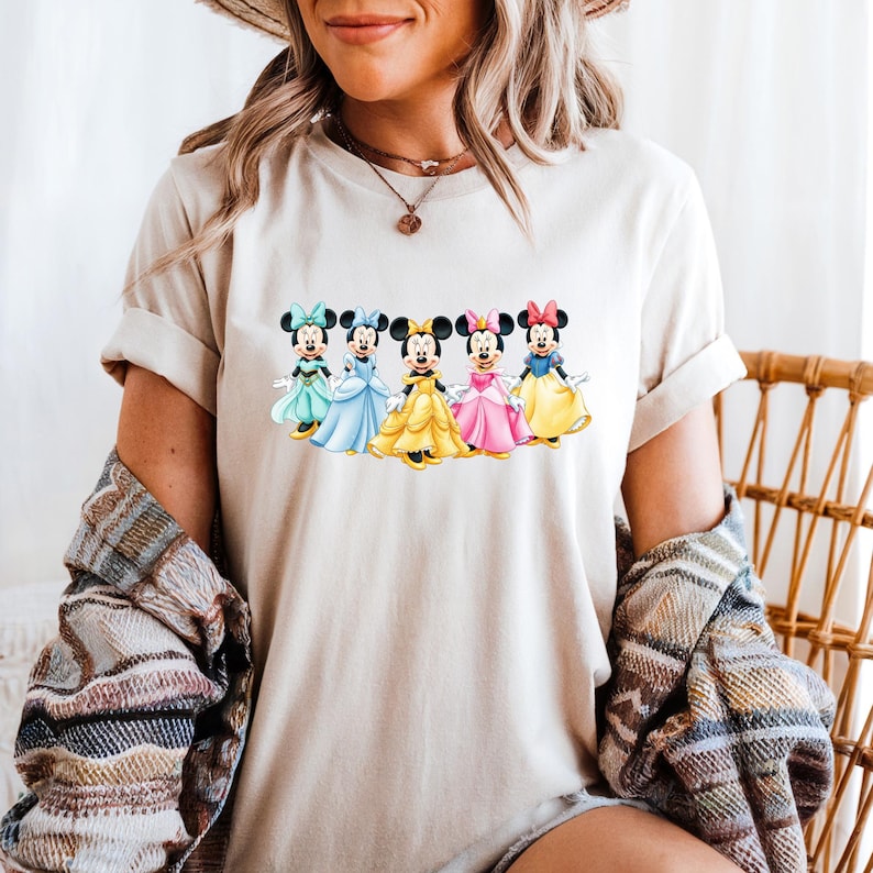 May include: A beige t-shirt featuring a colorful graphic of five Minnie Mouse characters in different dresses. Each Minnie Mouse is wearing a different colored dress, including teal, blue, yellow, pink, and yellow. The shirt is worn by a person.