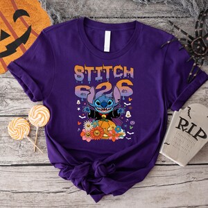 Puede incluir: Una camiseta morada con un dise&ntilde;o de Halloween de Stitch de Lilo & Stitch. El dise&ntilde;o incluye a Stitch disfrazado de vampiro, una calabaza, flores y el texto "STITCH 626".