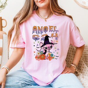 Puede incluir: Camiseta rosa con un personaje de dibujos animados con un sombrero de bruja, rodeado de flores e iconos de Halloween. La camiseta muestra el texto "ANGEL 624" en una fuente colorida y divertida. Es una camiseta de manga corta y cuello redondo.