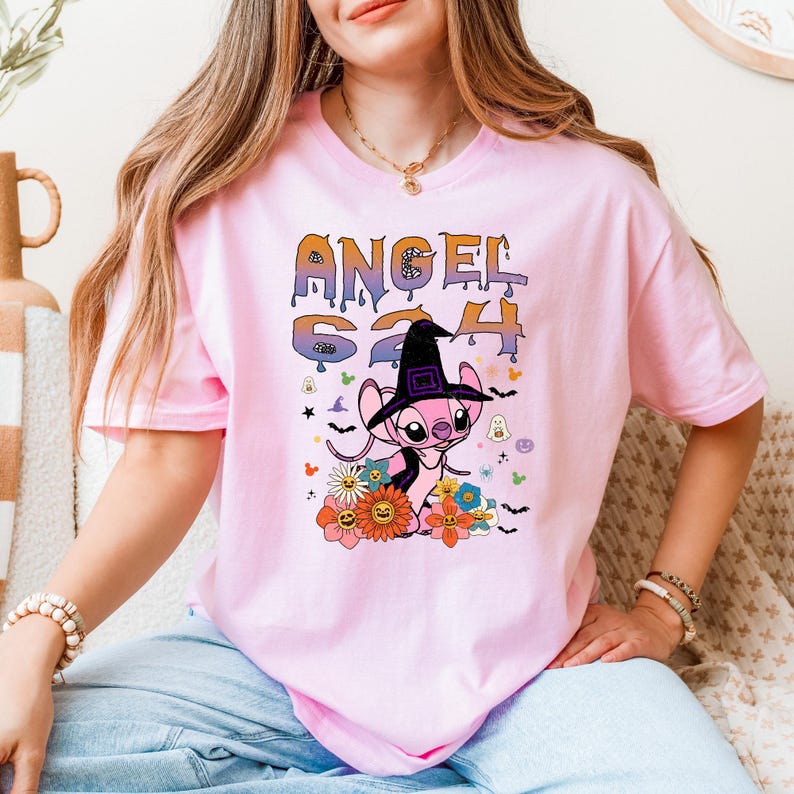 Puede incluir: Camiseta rosa con un personaje de dibujos animados con un sombrero de bruja, rodeado de flores e iconos de Halloween. La camiseta muestra el texto "ANGEL 624" en una fuente colorida y divertida. Es una camiseta de manga corta y cuello redondo.