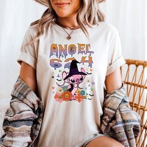 Puede incluir: Camiseta color crema con un gr&aacute;fico de un personaje alien&iacute;gena rosa con un sombrero de bruja, rodeado de flores y el texto "ANGEL 624". El dise&ntilde;o tiene un tema de Halloween con fantasmas y murci&eacute;lagos.