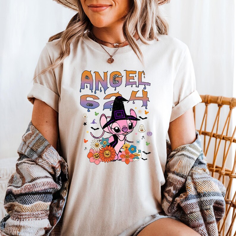 Puede incluir: Camiseta color crema con un gr&aacute;fico de un personaje alien&iacute;gena rosa con un sombrero de bruja, rodeado de flores y el texto "ANGEL 624". El dise&ntilde;o tiene un tema de Halloween con fantasmas y murci&eacute;lagos.