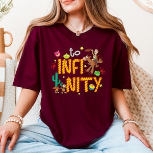 May include: Burgundy t-shirt with a graphic of characters from the Toy Story movie franchise. The graphic includes Woody riding Bullseye, Buzz Lightyear, and other characters. The text on the shirt reads "to infinity and beyond".