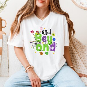 May include: White t-shirt with a green and purple graphic that says "And Beyond" with a cartoon rocket, space characters, and stars.