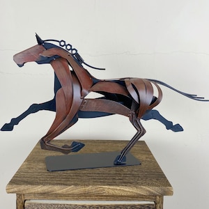 May include: A metal sculpture of a running horse. The horse is crafted from brown and black metal pieces, with a flowing mane and tail. The sculpture is mounted on a black base and sits on a wooden surface.
